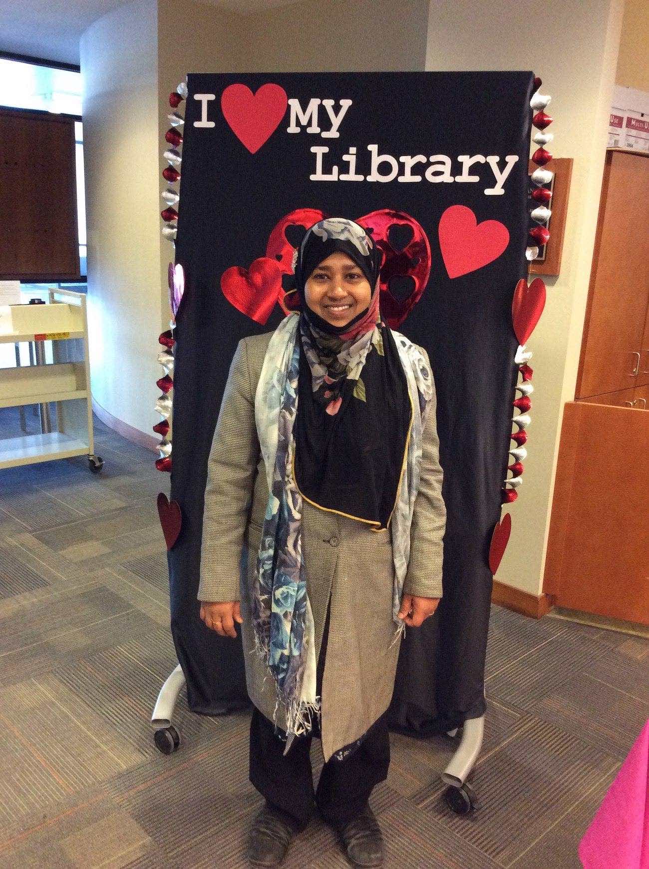 a smiling student in a black and white patterned hijab stands in front of the I love my library backdrop.